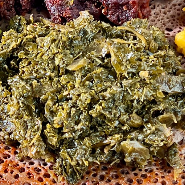 Close-up of green Ethiopian collard greens (gomen) on injera bread, with other dishes blurred.