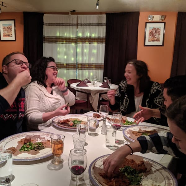 Friends enjoying Ethiopian food and drinks at Bete-Lukas restaurant, laughing and eating with their hands.