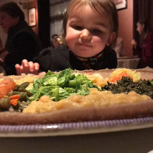 A child peers over a large platter of colorful Ethiopian dishes, including injera, vegetables, and stews.